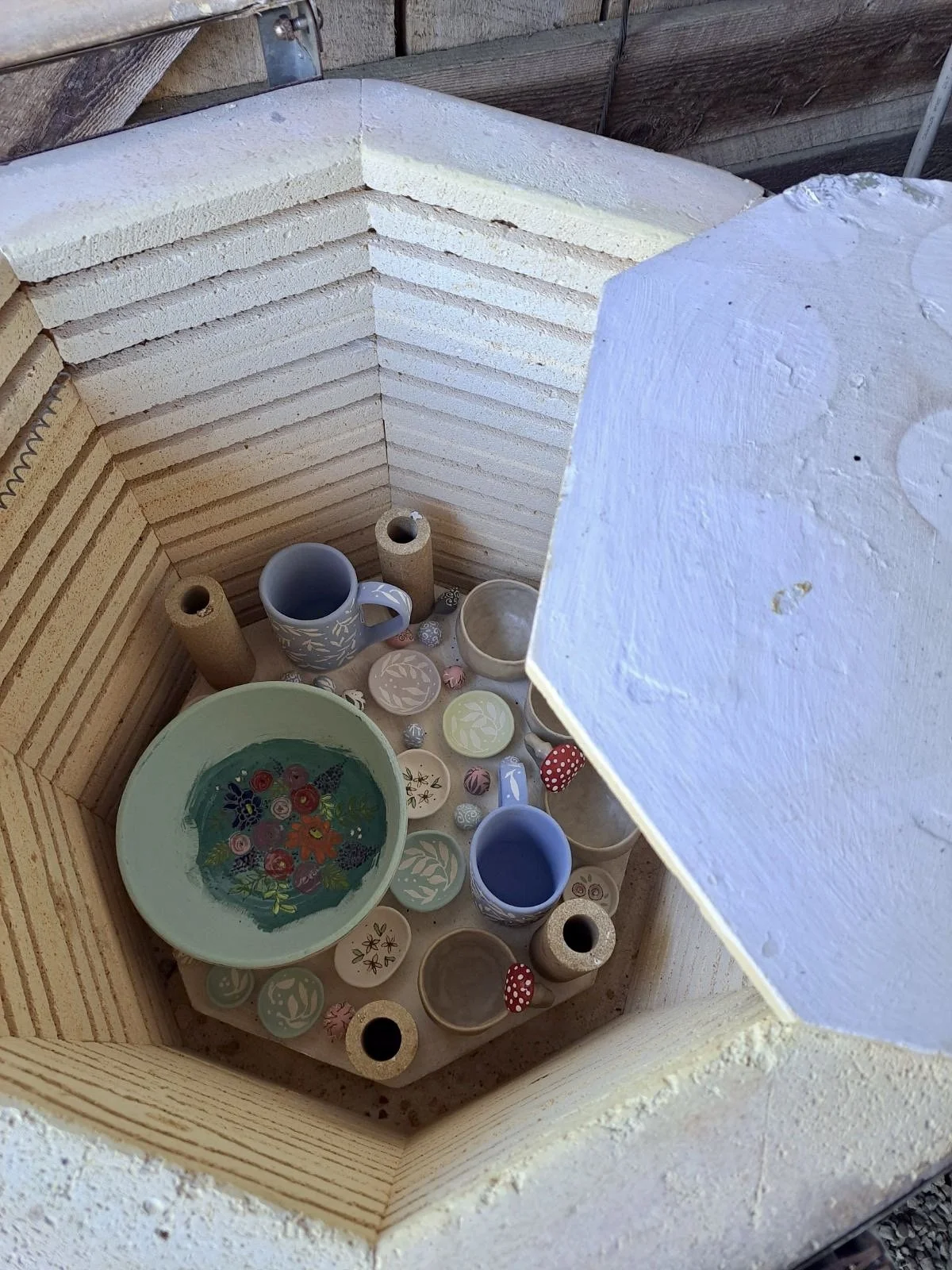 Inside a wooden outdoor kiln with various ceramics including cups, bowls, and small decorative items.