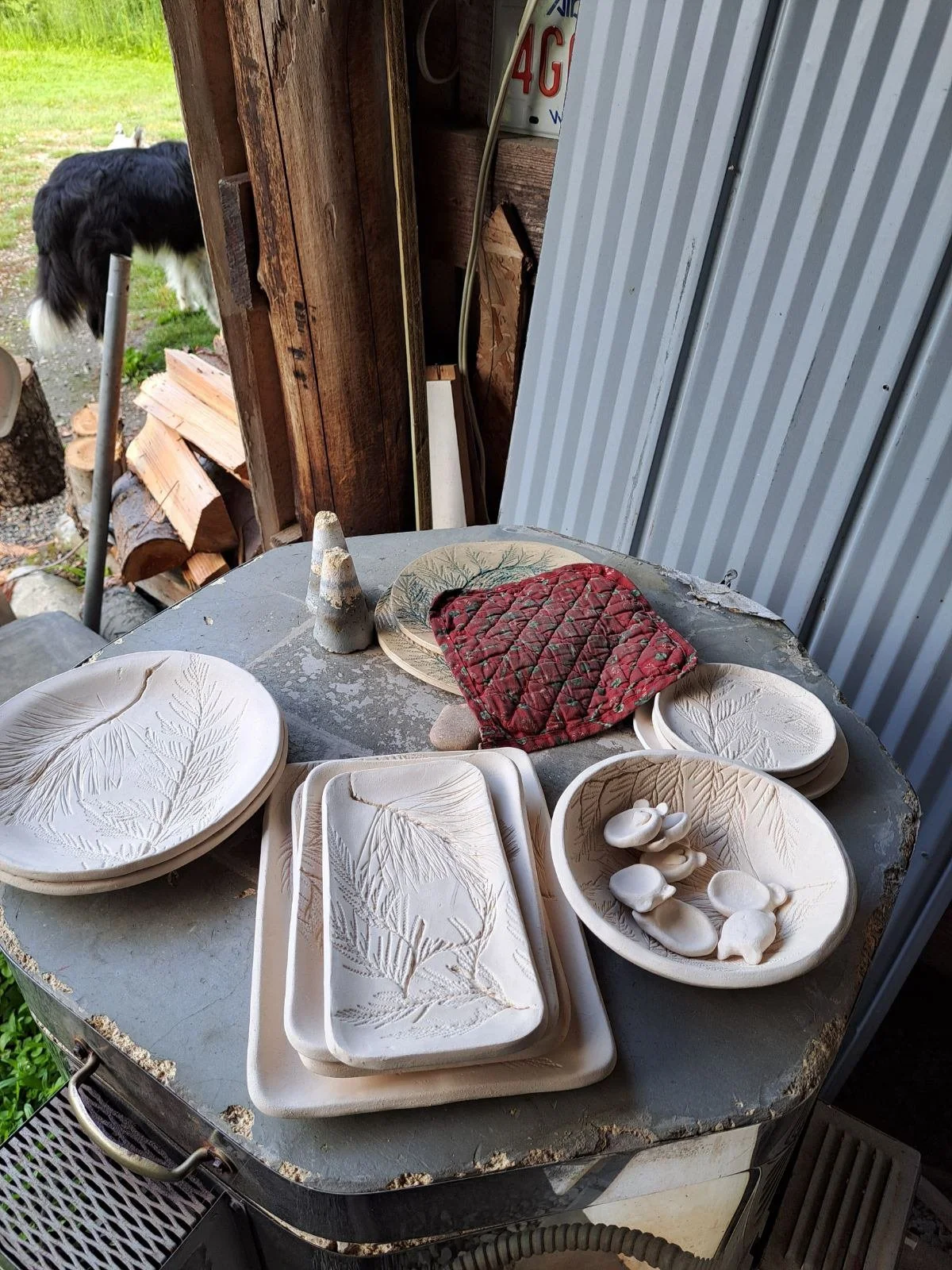 A table with various unglazed ceramic pottery items placed on it, including plates, trays, a bowl with ceramic stones, and a small cone. A red quilted oven mitt is also on the table. In the background, part of a corrugated metal wall and a wooden wal