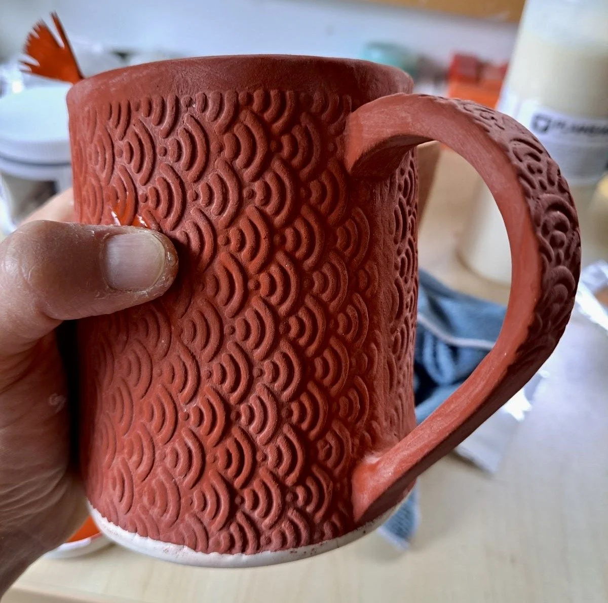 A person's hand holding a red ceramic mug with raised wave-like patterns and an ornate handle.