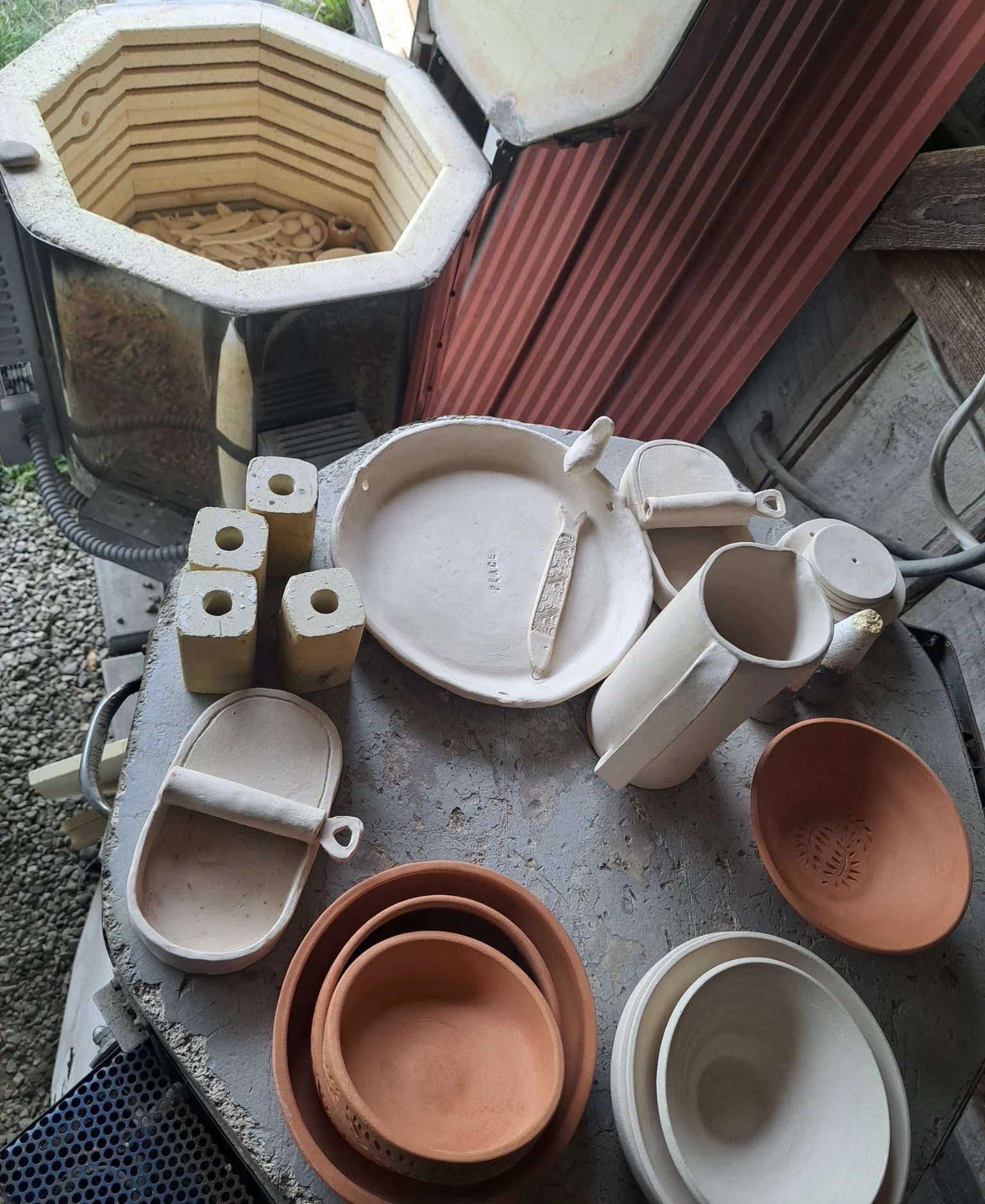 Unfinished ceramic pottery and clay bowls on a worktable, with a kiln in the background.