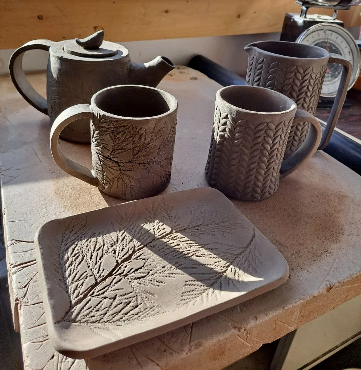 Set of handcrafted ceramic teapot, two mugs, and a tray with leaf patterns on a wooden table.