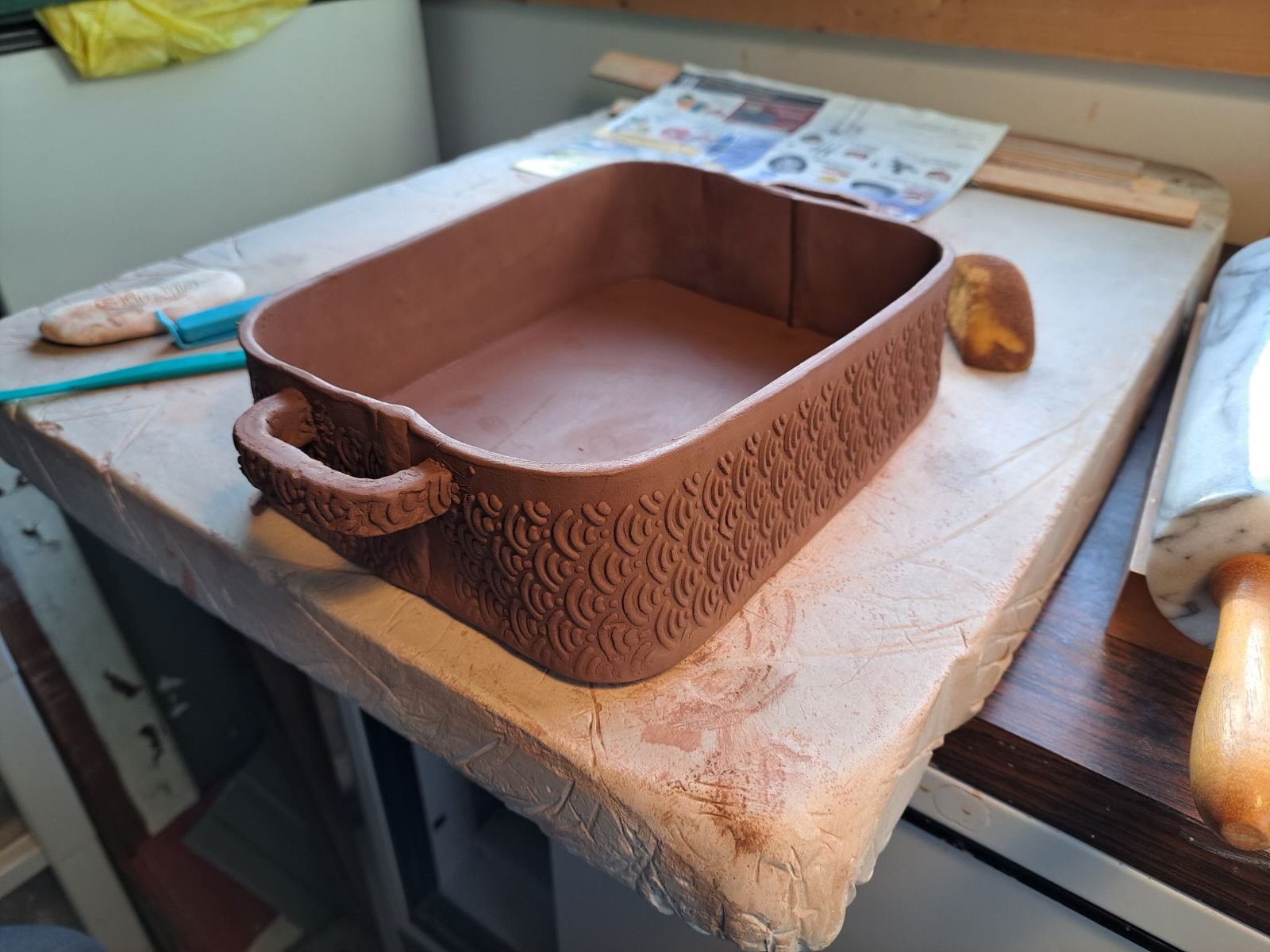 Unfinished rectangular ceramic casserole dish with decorative pattern, resting on a worktable, surrounded by tools and clay.