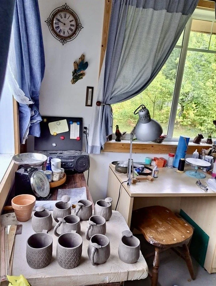 Cluttered craftsman's workspace with pottery tiles, handmade ceramic mugs, a clock, and a window with a view of green trees.