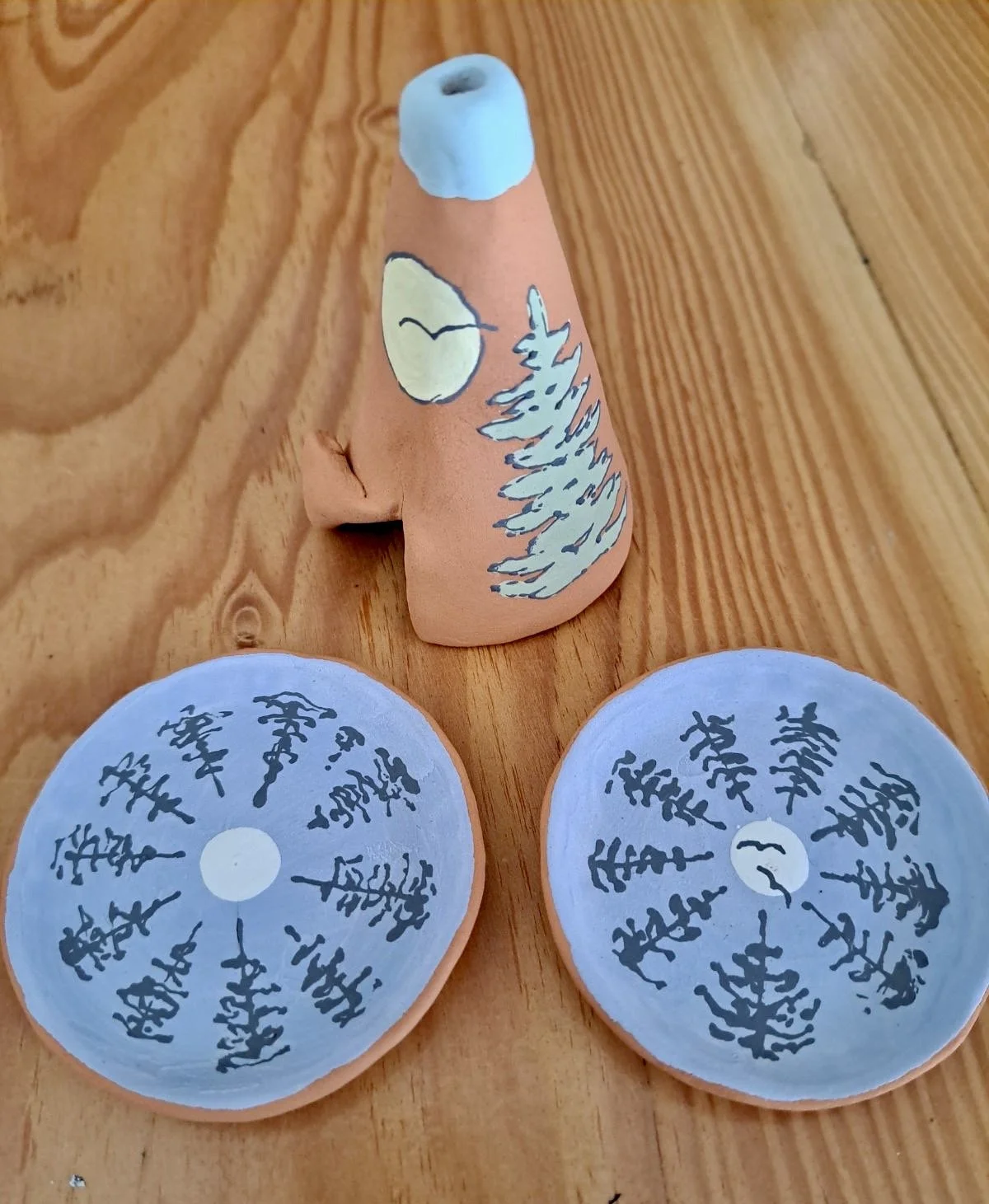 Clay sculptures of a moon and pine trees, with a face on the cone-shaped sculpture, and painted tree designs on two circular objects, placed on a wooden surface.