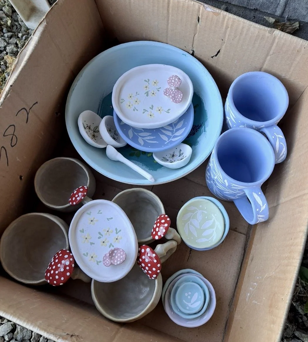 A box containing various ceramic tea set pieces, including cups, bowls, plates, and a spoon, with some decorated with floral and mushroom patterns in pastel colors.