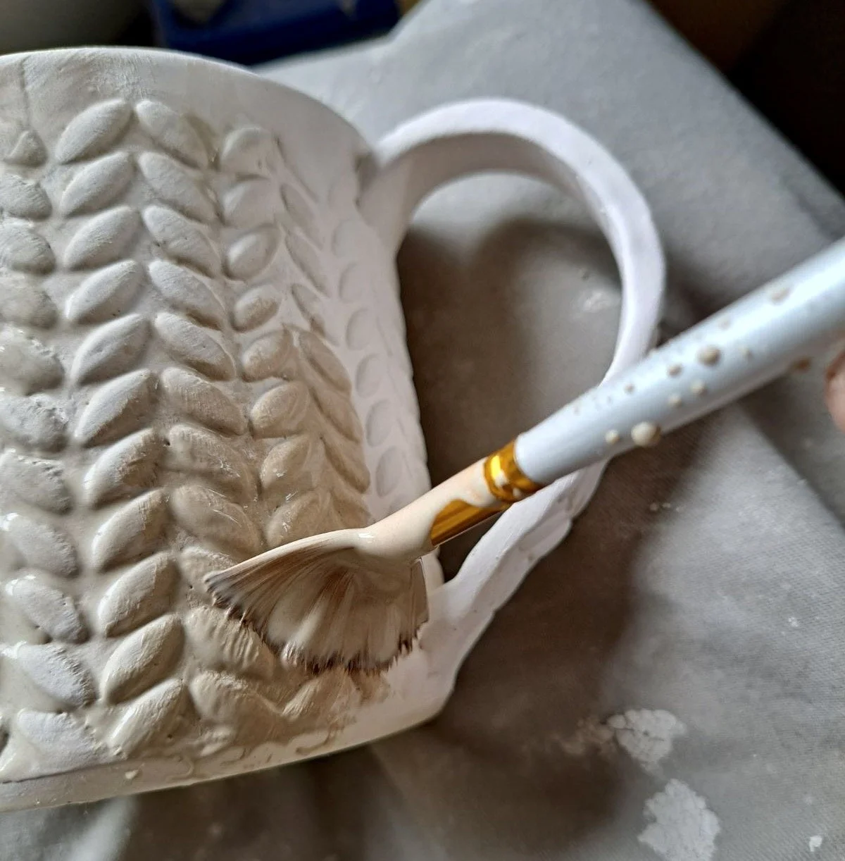 A ceramic mug with a textured surface featuring leaf-like patterns, being decorated with a paintbrush dipped in brown paint.