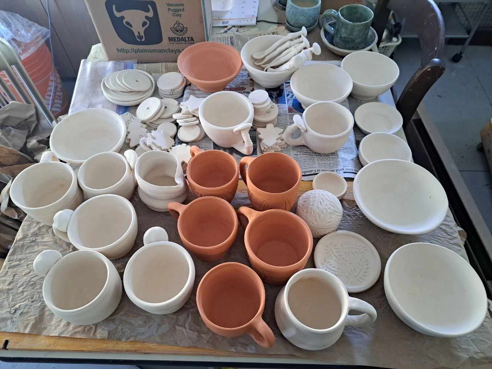 A table covered with unglazed ceramic pottery pieces including cups, bowls, and decorative items, some in plain white and others in terracotta color, with tools and materials for pottery at the background.
