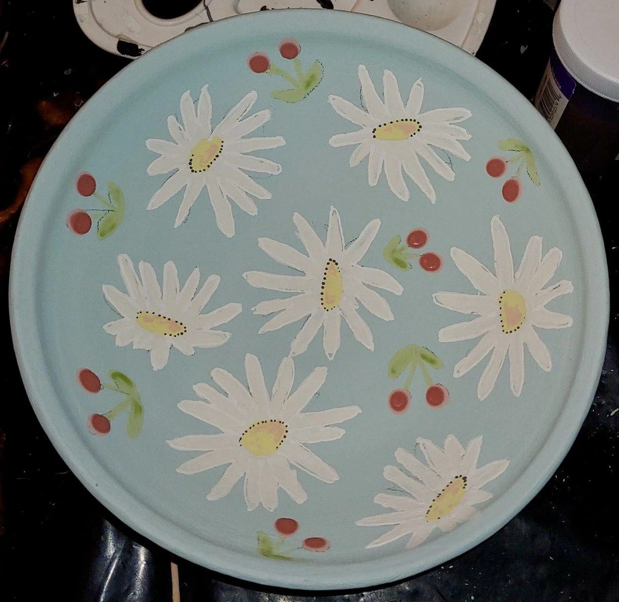 A painted round tray with a light blue background featuring large white daisies with yellow centers and small red berries with green leaves.
