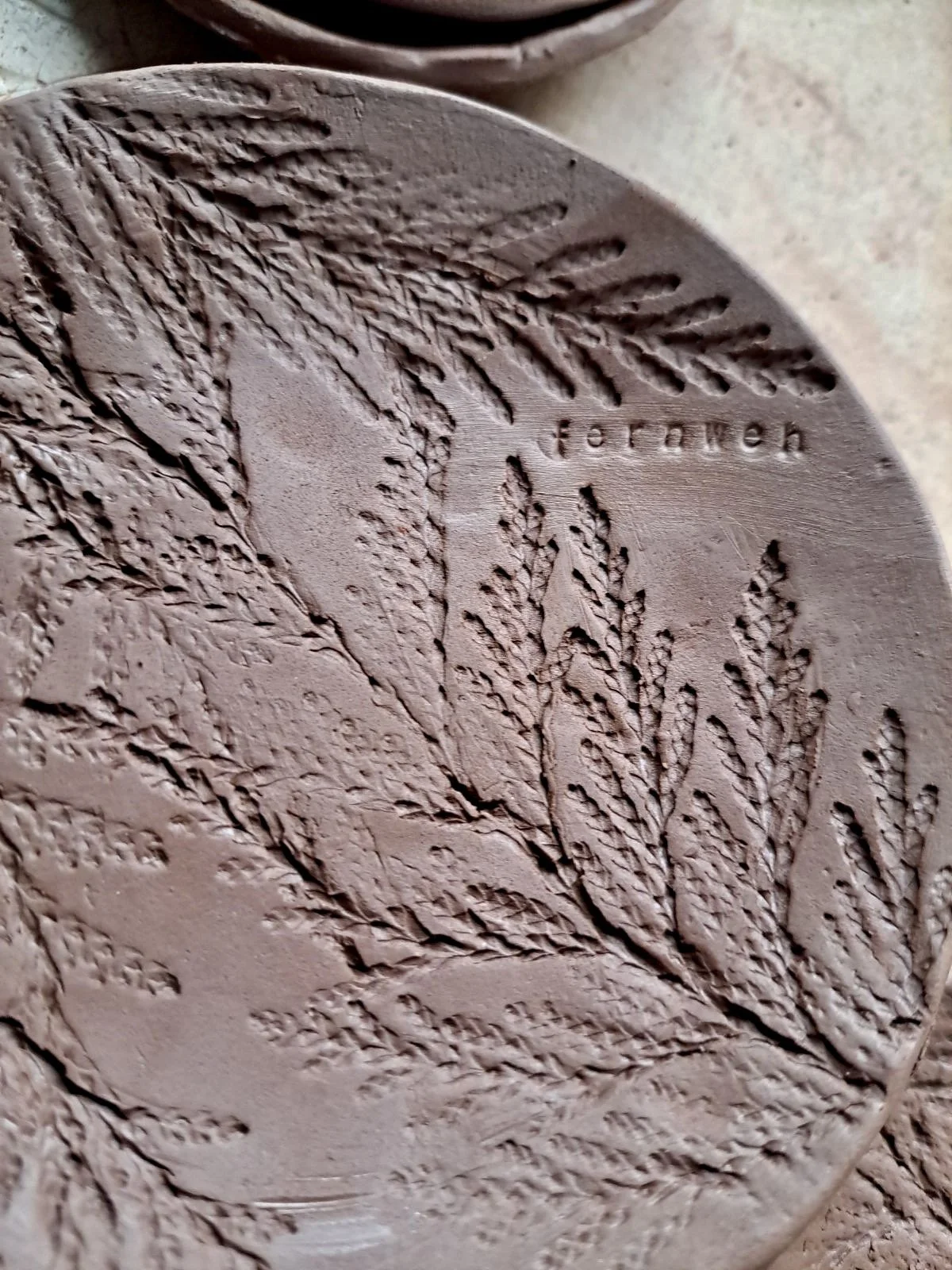 A round clay stamp with embossed fern and leaf patterns, and the word 'fern' embossed on it.