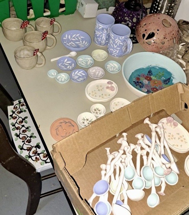A table displays various handmade ceramic items including cups, bowls, and plates, along with a cardboard box filled with ceramic spoons and some decorative pieces, in a setting that appears to be a craft or thrift store.