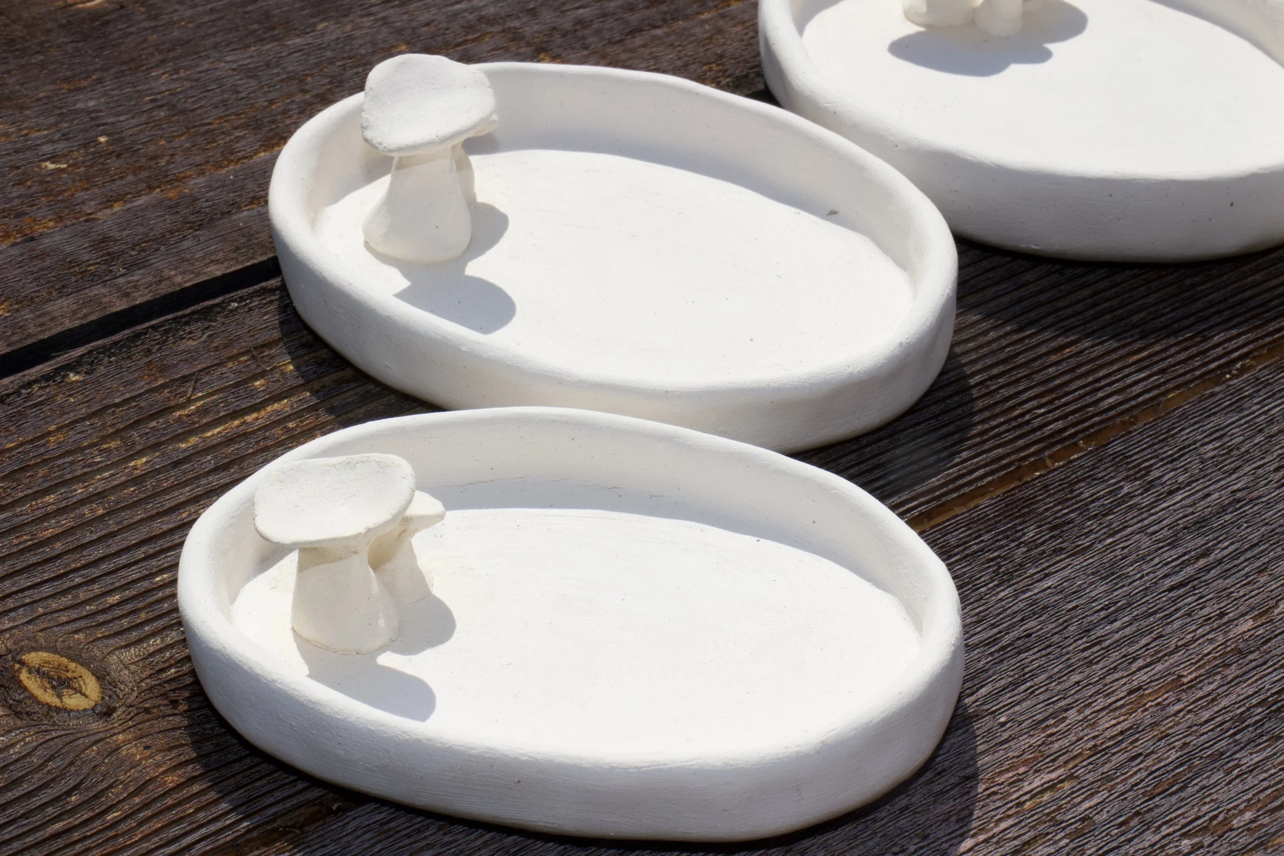 Three white ceramic dishes with small mushroom sculptures on a dark wooden surface.