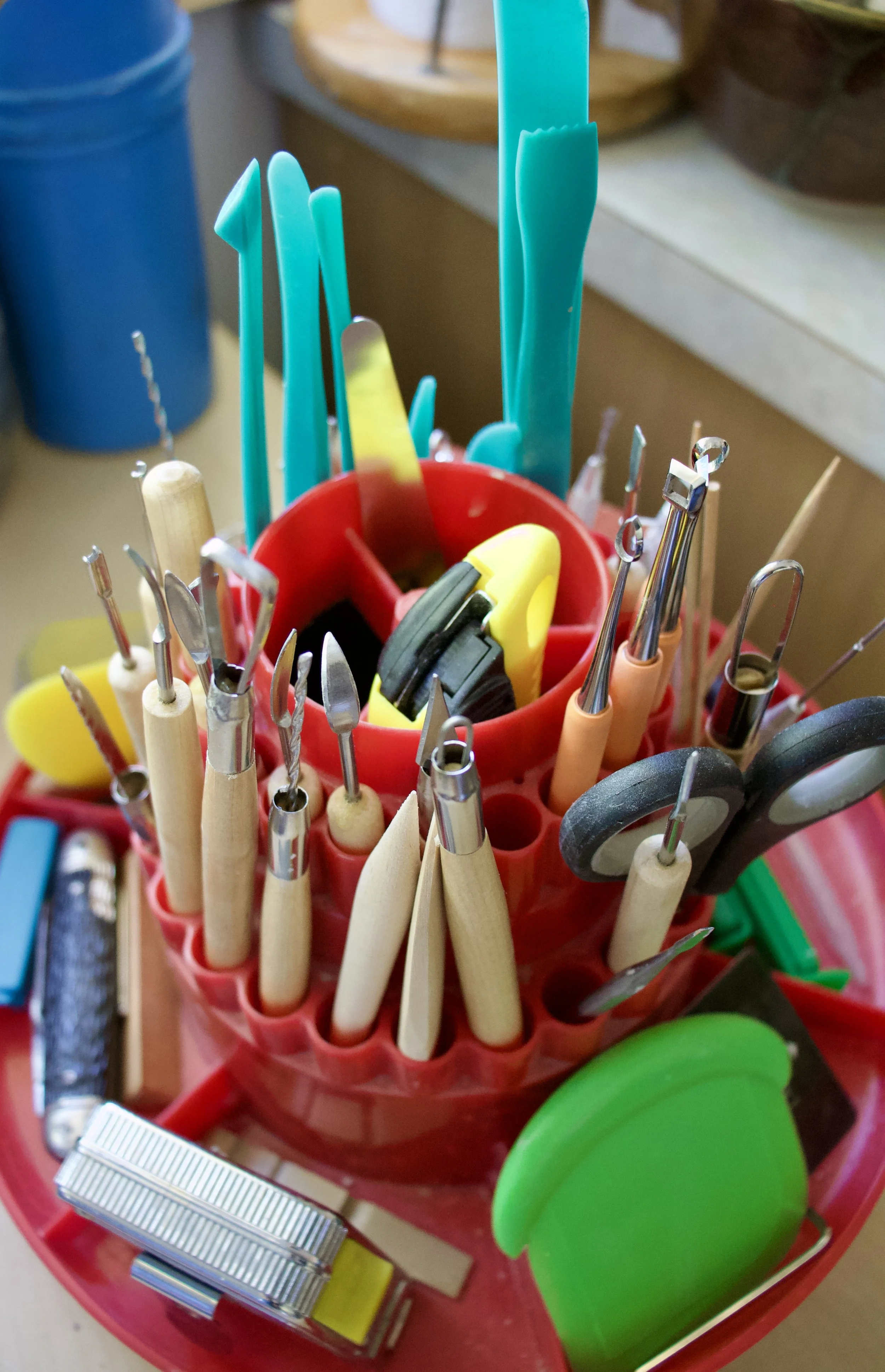 A collection of various hand tools, including scalpels, cutters, and scissors, neatly organized in a red multi-compartment container, with some tools stored in a green container and a yellow tape measure at the base.