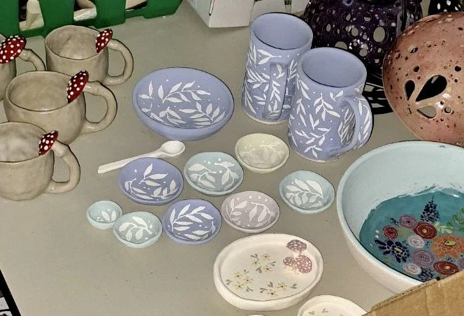 A collection of ceramic mugs, bowls, and small dishes, some with blue and white leaf patterns, some plain, with a large bowl containing colorful decorated paper flowers and a pink decorative item nearby.