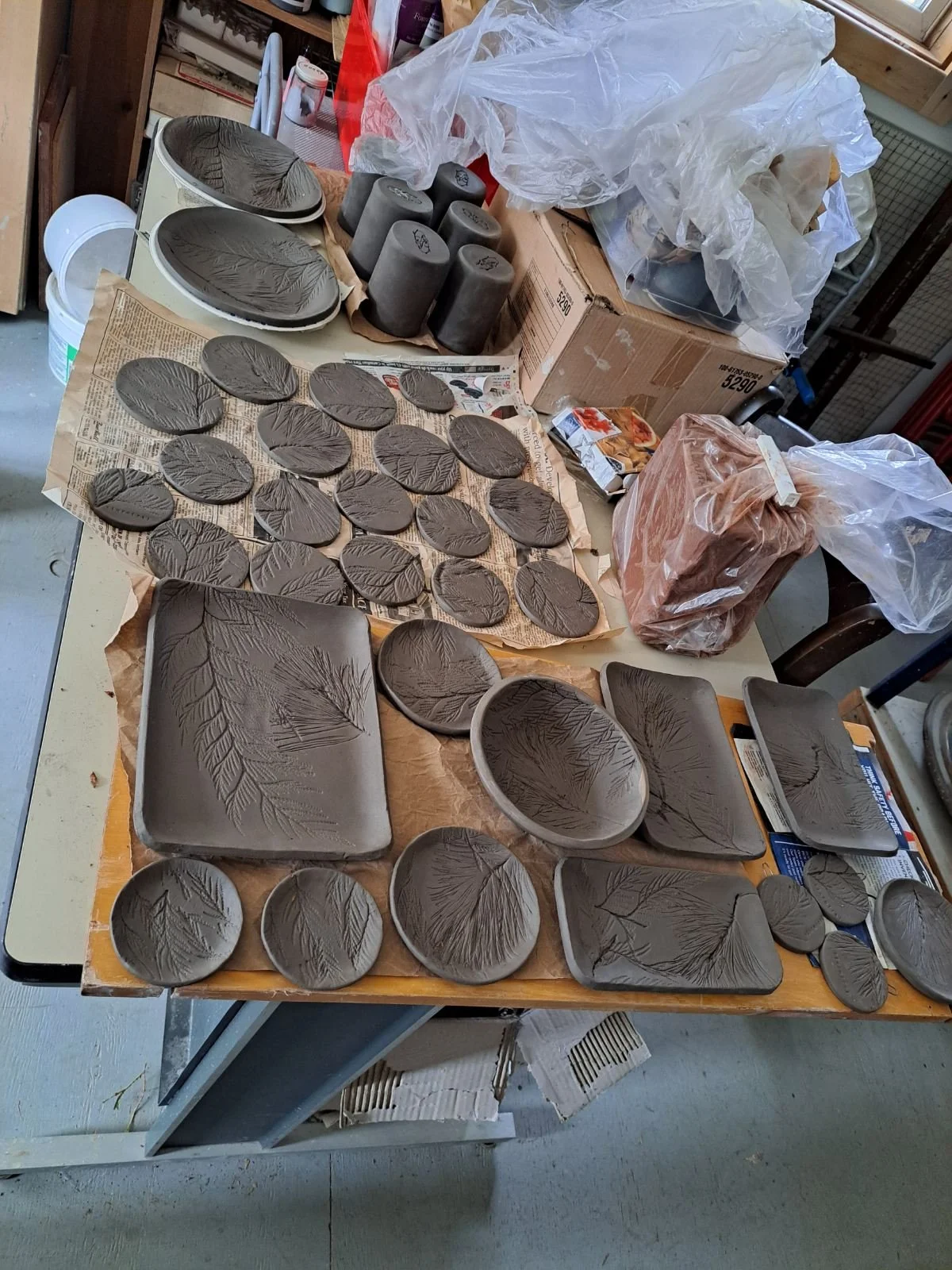 Clay pottery pieces with leaf patterns drying on newspaper and trays in a pottery studio.