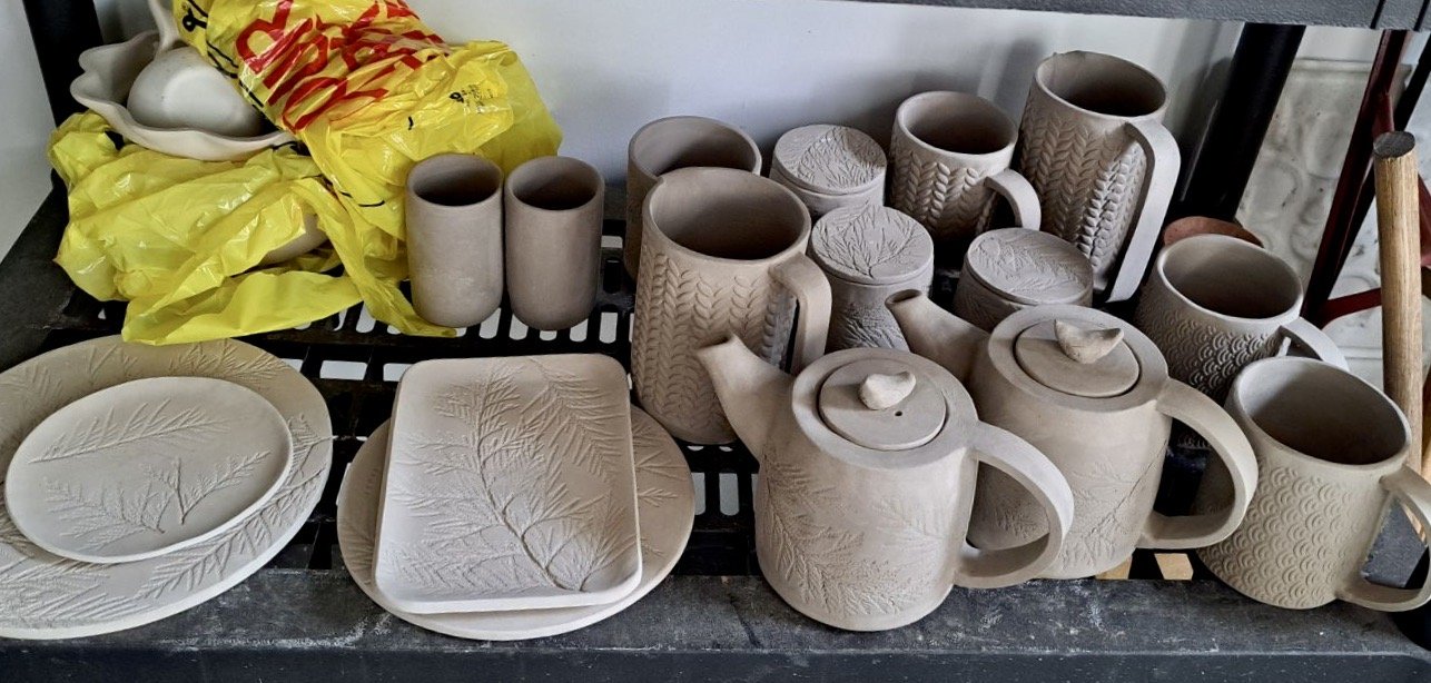Unfinished clay pottery including mugs, teapots, plates, and bowls with etched leaf and fern patterns, placed on a black surface with yellow shopping bags in the background.