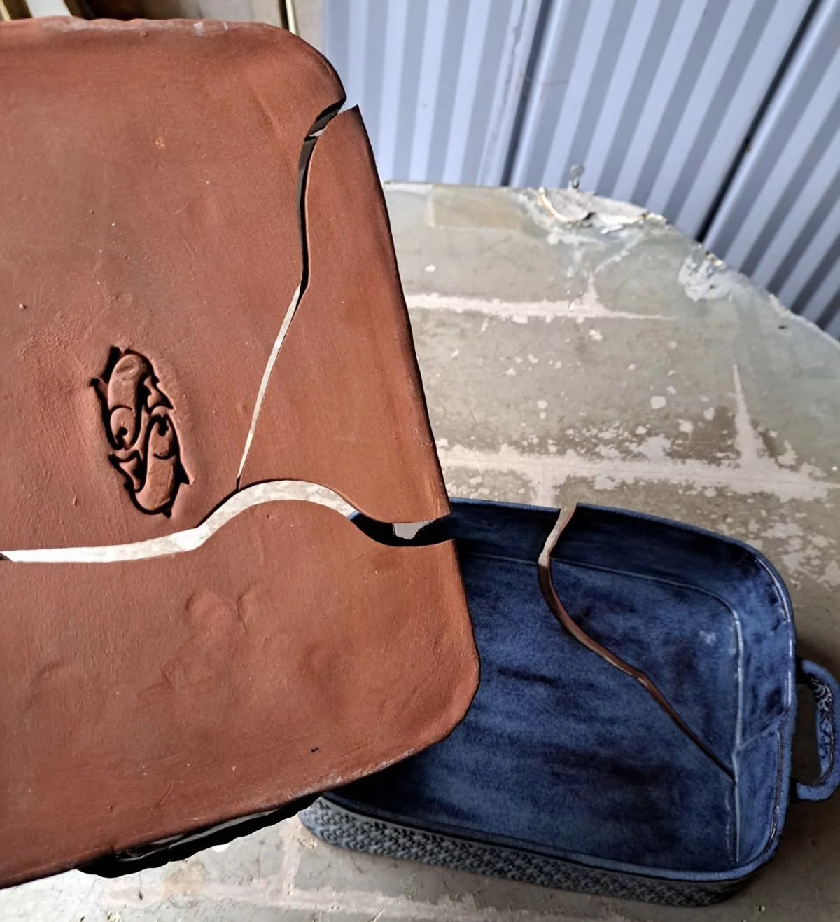 Broken brown leather chair with a logo, placed on top of a blue hard-shell suitcase, on a concrete floor with a brick and metal wall in the background.