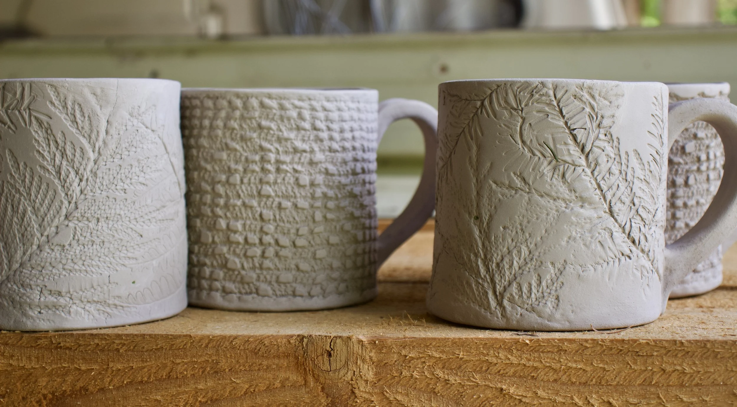 Three white ceramic mugs with raised leaf and fern patterns, placed on a wooden surface.