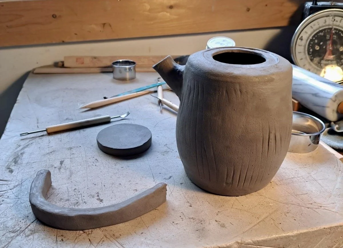 Clay teapot on worktable with pottery tools, a scale, and a bowl in the background.