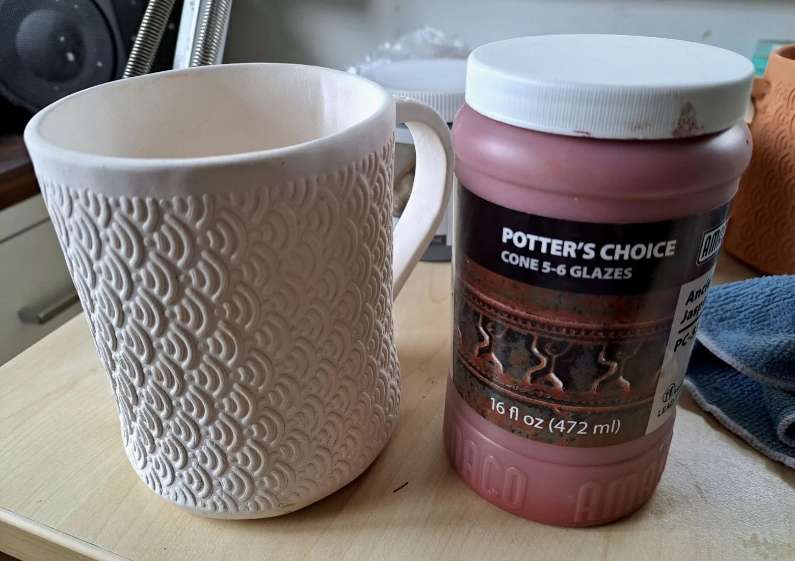 A white coffee mug with textured patterns next to a pink container of Potter's Choice cone 5-6 glazes on a light-colored table.