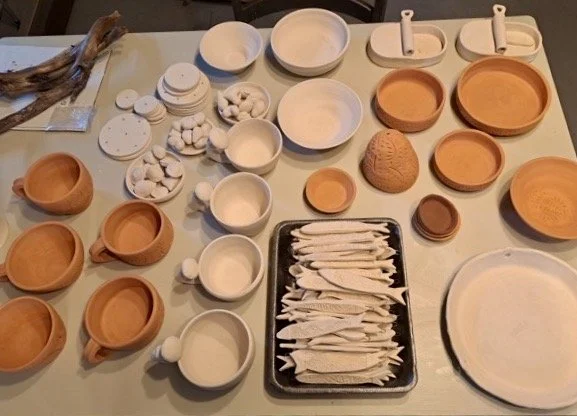 A table with various unglazed clay bowls, plates, and cups, some with white objects and others empty, along with a tray of stacked white cloth napkins or towels, and a large round piece of textured clay.