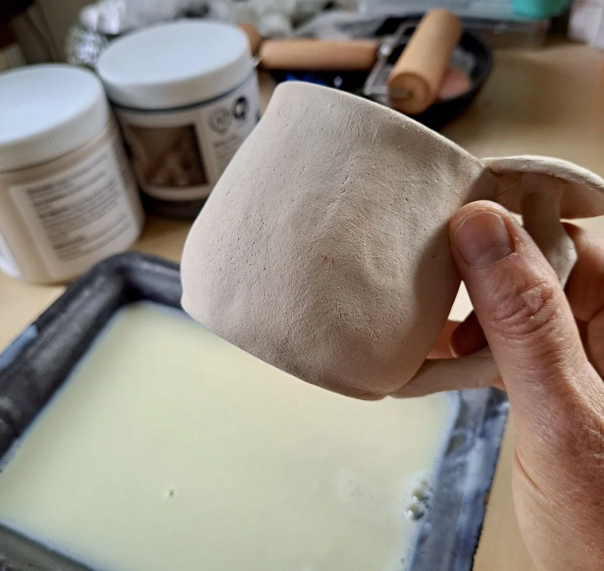 A person holding a ceramic cup with a textured, unglazed surface above a tray of liquid ceramic slip. In the background, there are containers of clay or similar materials and pottery tools on a worktable.