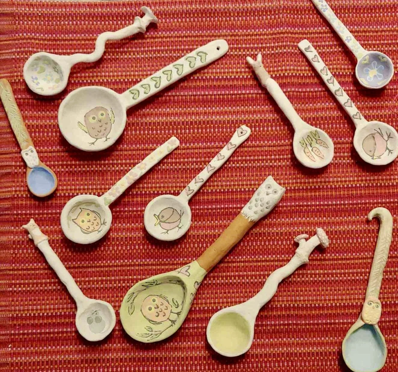 Set of decorative ceramic spoons and utensils with hand-painted animal and nature illustrations on each, arranged on a red woven fabric background.