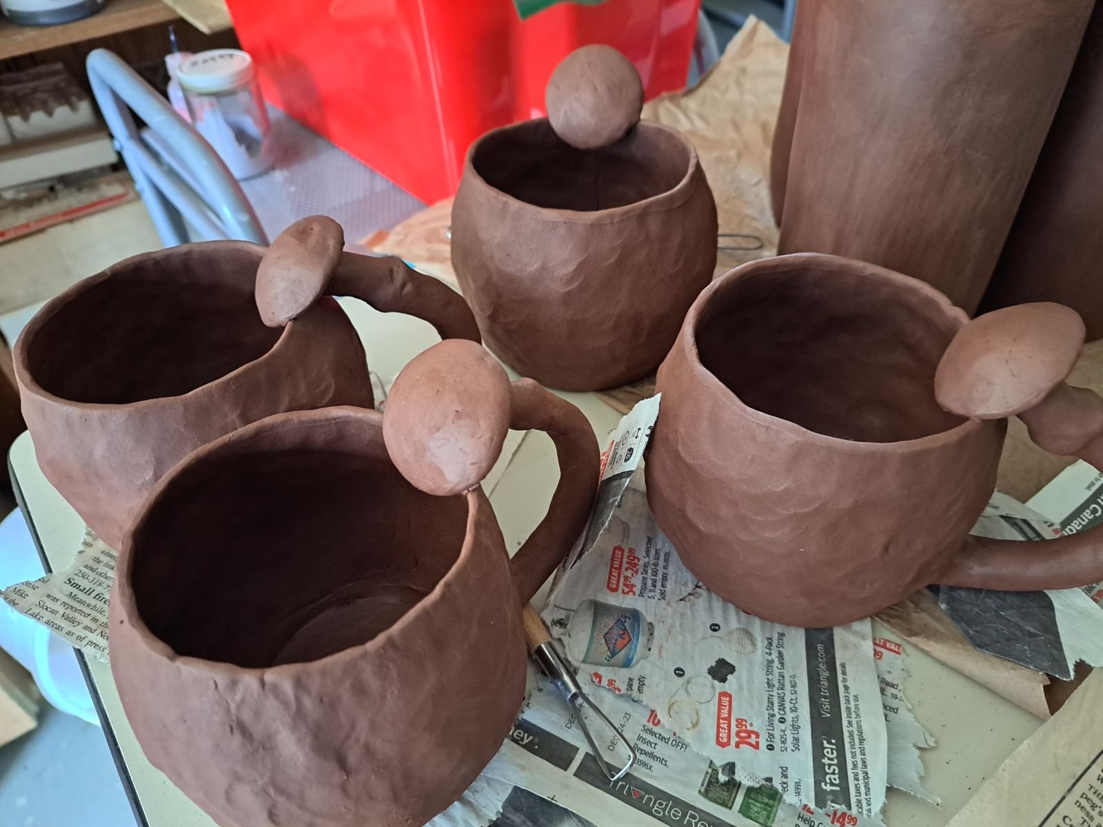 Four unfinished ceramic cups with handles and spherical lids in a workshop, placed on newspaper-covered surface.