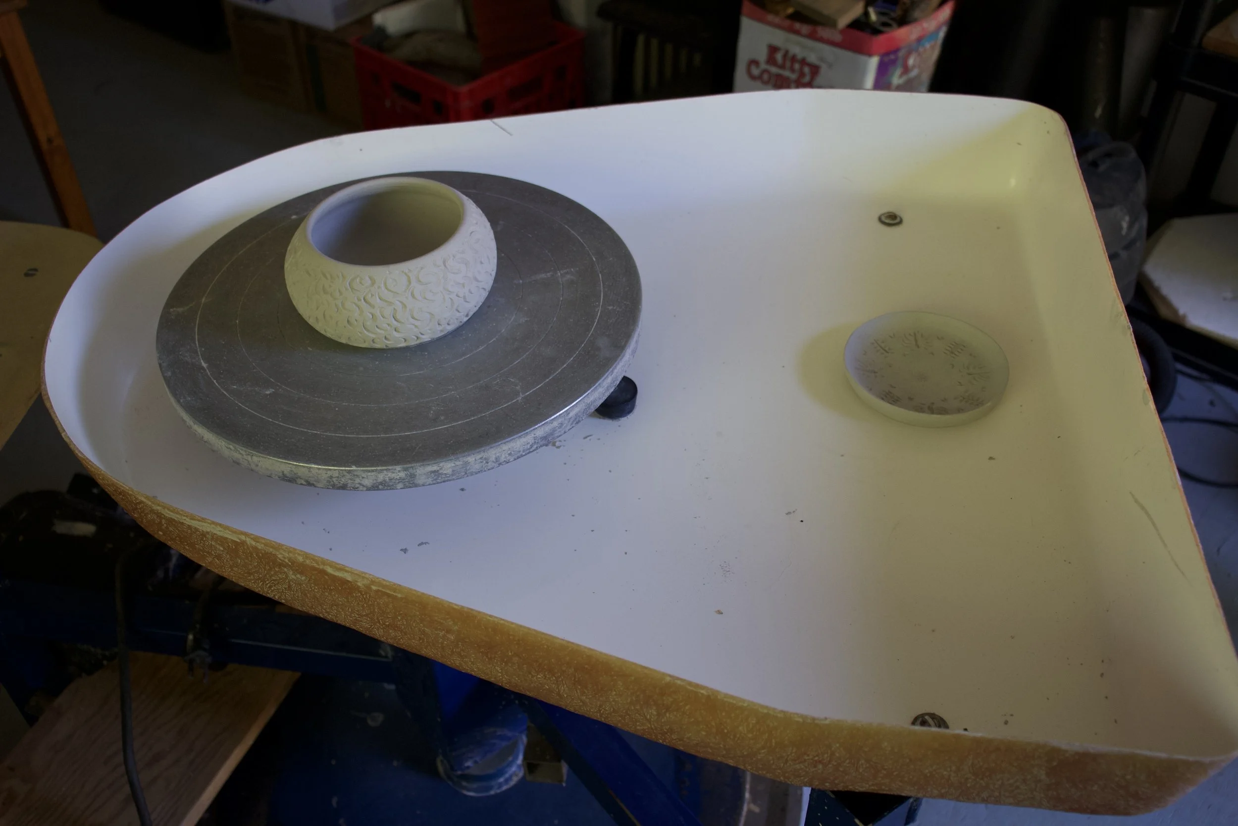 A ceramic bowl on a pottery wheel in a workshop. There is also a small plate on the right side of the wheel. The workshop has tools and storage in the background.