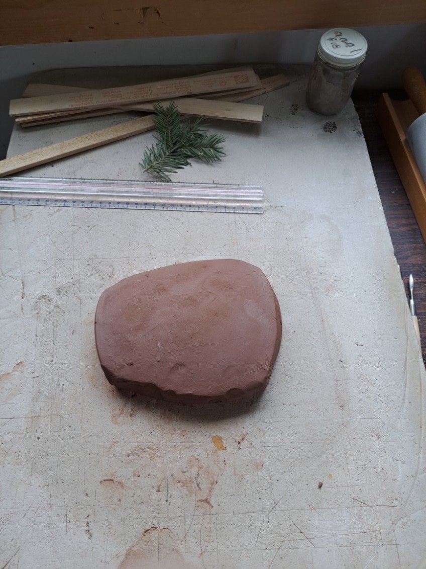 A piece of red clay on a worktable with gardening sprigs, a ruler, wooden sticks, a jar of powder, and carving tools.