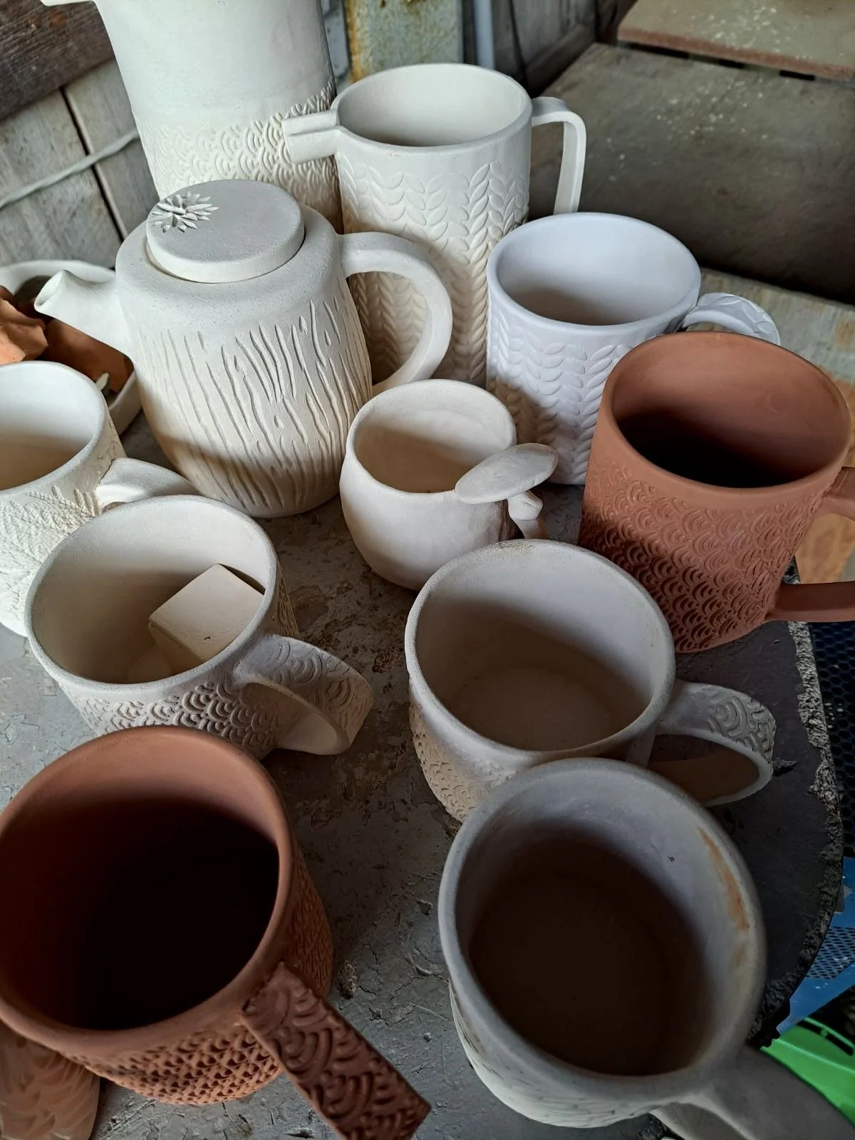 Various ceramic mugs and teapots with textured patterns on a rustic surface, some filled with dark liquid, in a rustic setting.