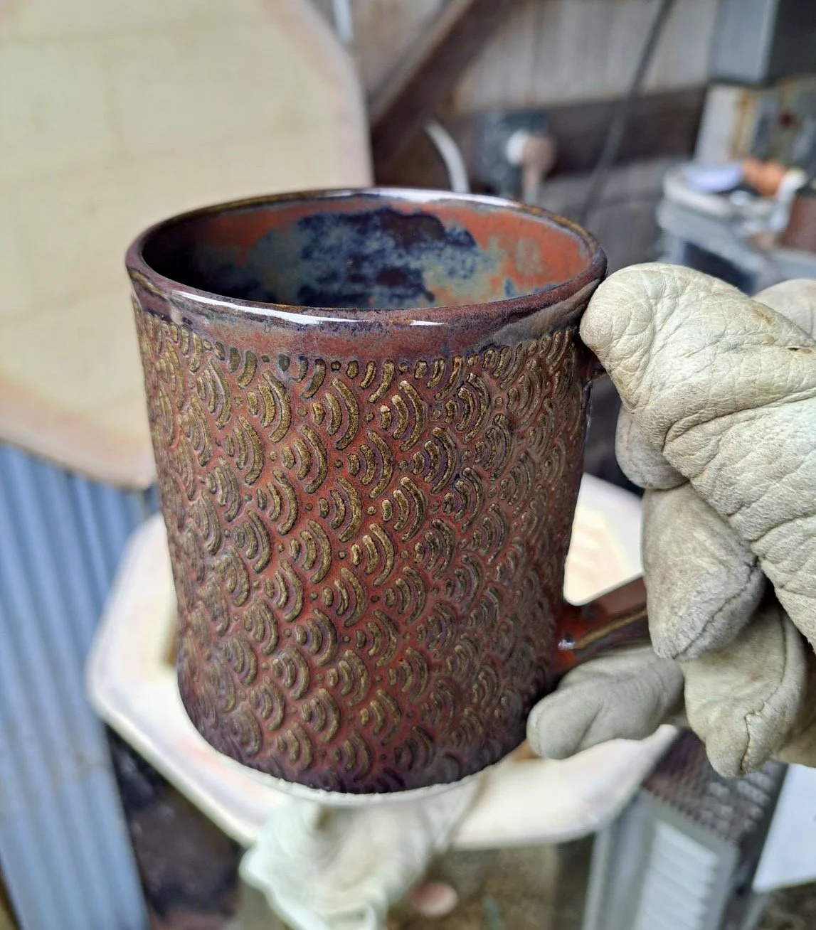 A person wearing a glove holding a textured ceramic mug with a reddish-brown glaze and intricate raised patterns, with a workshop background.