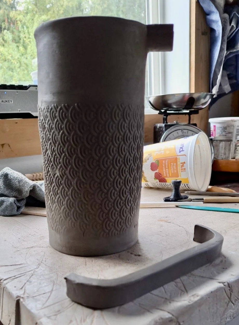 A tall, cylindrical ceramic mug with a textured pattern, placed on a workbench with pottery tools and pottery supplies in the background.