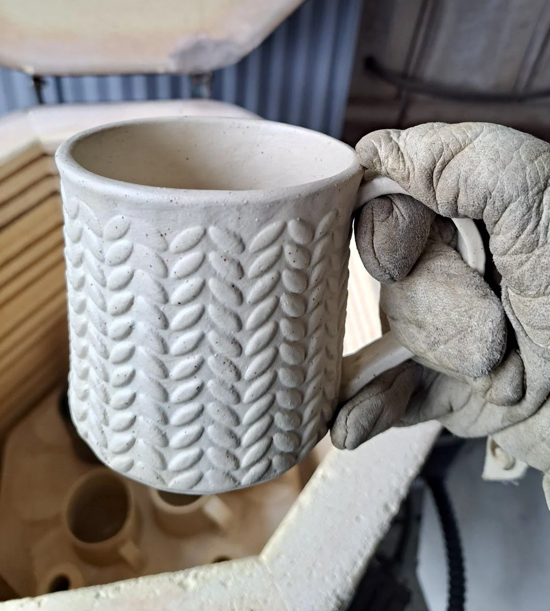 A person wearing a dusty, work glove holds a white ceramic mug with a textured vine pattern in a pottery studio. The background shows pottery molds and shelves.