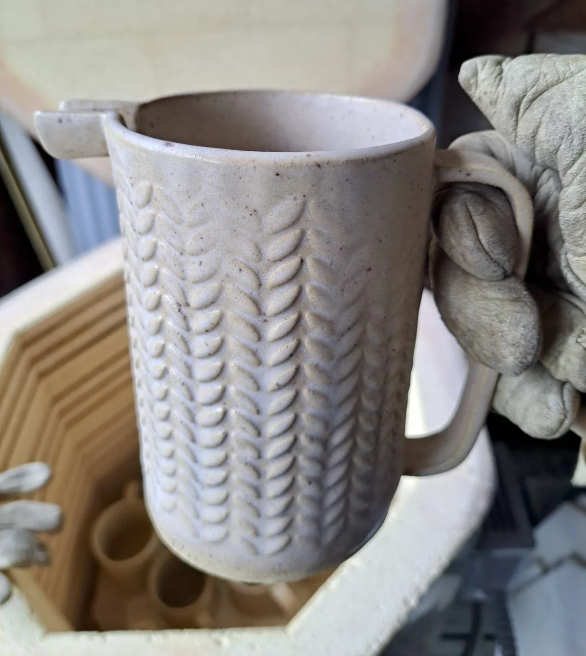 A ceramic mug with a textured wheat pattern, held by a gloved hand in a pottery studio.