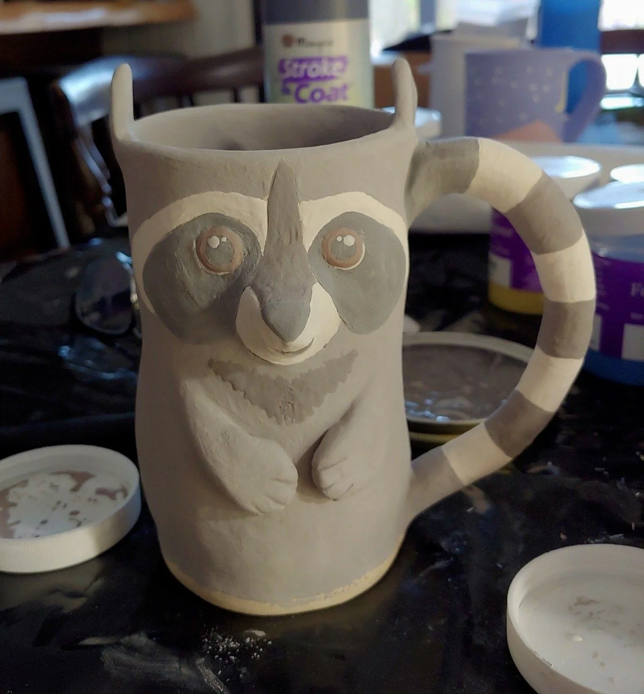 A ceramic mug featuring a raccoon's face, with painted eyes, nose, and mouth, and striped pattern on its tail handle, set on a black surface surrounded by small open containers of paint or glaze.