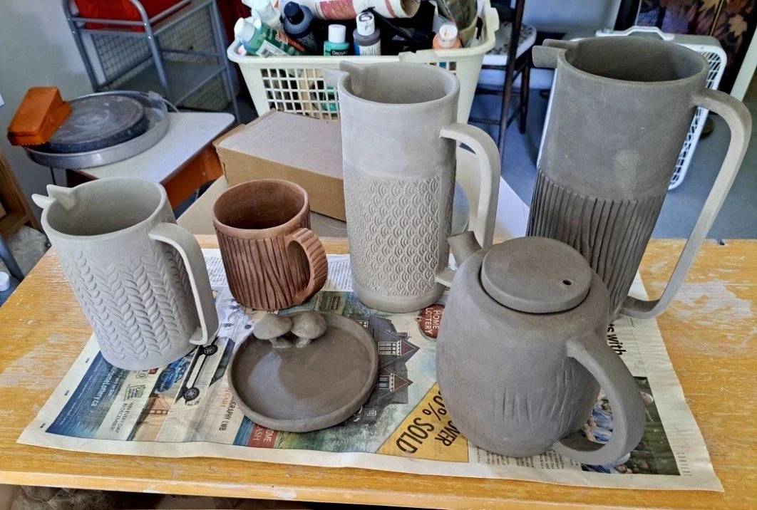 Unfinished ceramic mugs and plates on a table covered with newspaper, with various pottery tools and supplies in the background.