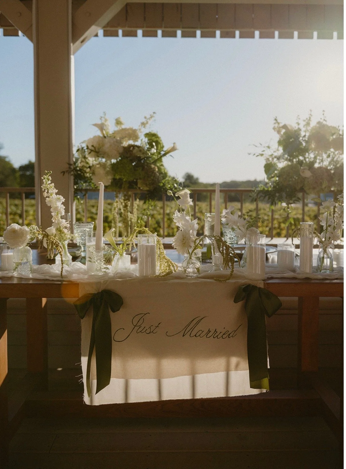 A decorated table with a white banner reading 'Just Married,' black ribbons, and floral arrangements set outdoors in a pavilion with a view of a field for a wedding celebration.