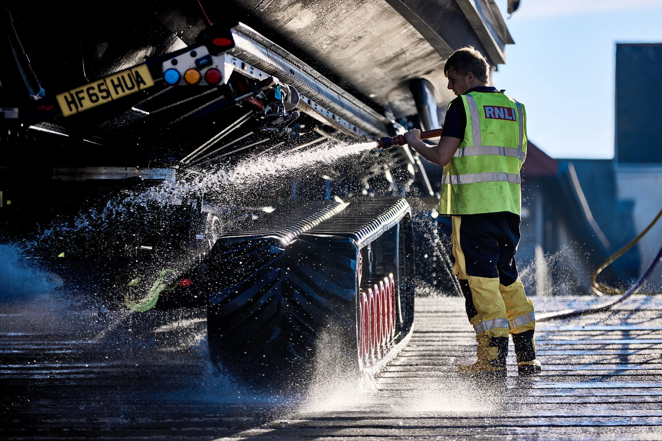 A person wearing a high-visibility vest with RNLI logo washing a boat with a hose, water splashing around in an outdoor setting.