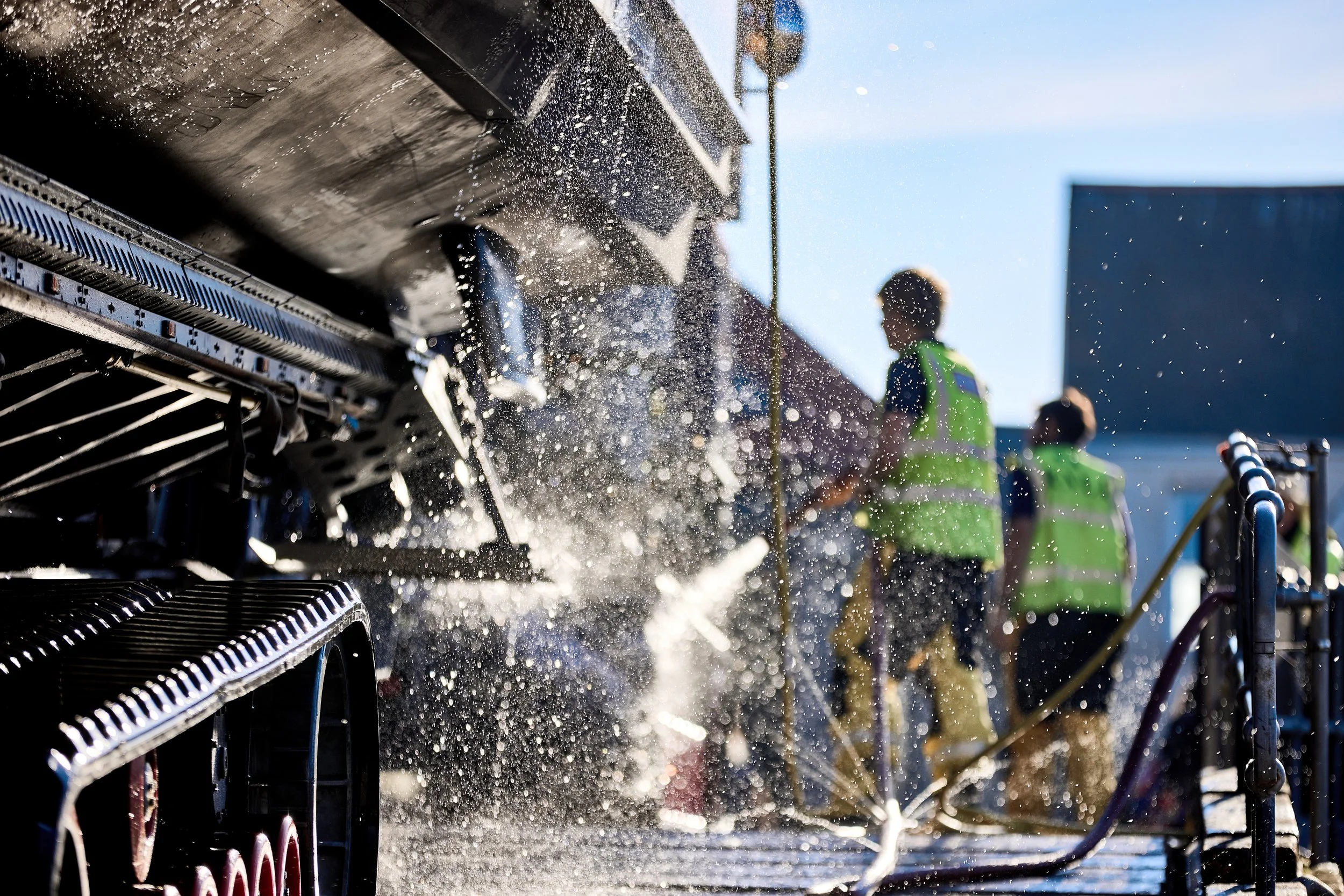 Close-up of a side of a snowblower being used to clear snow, with snow spraying out and people in the background wearing neon green safety vests.