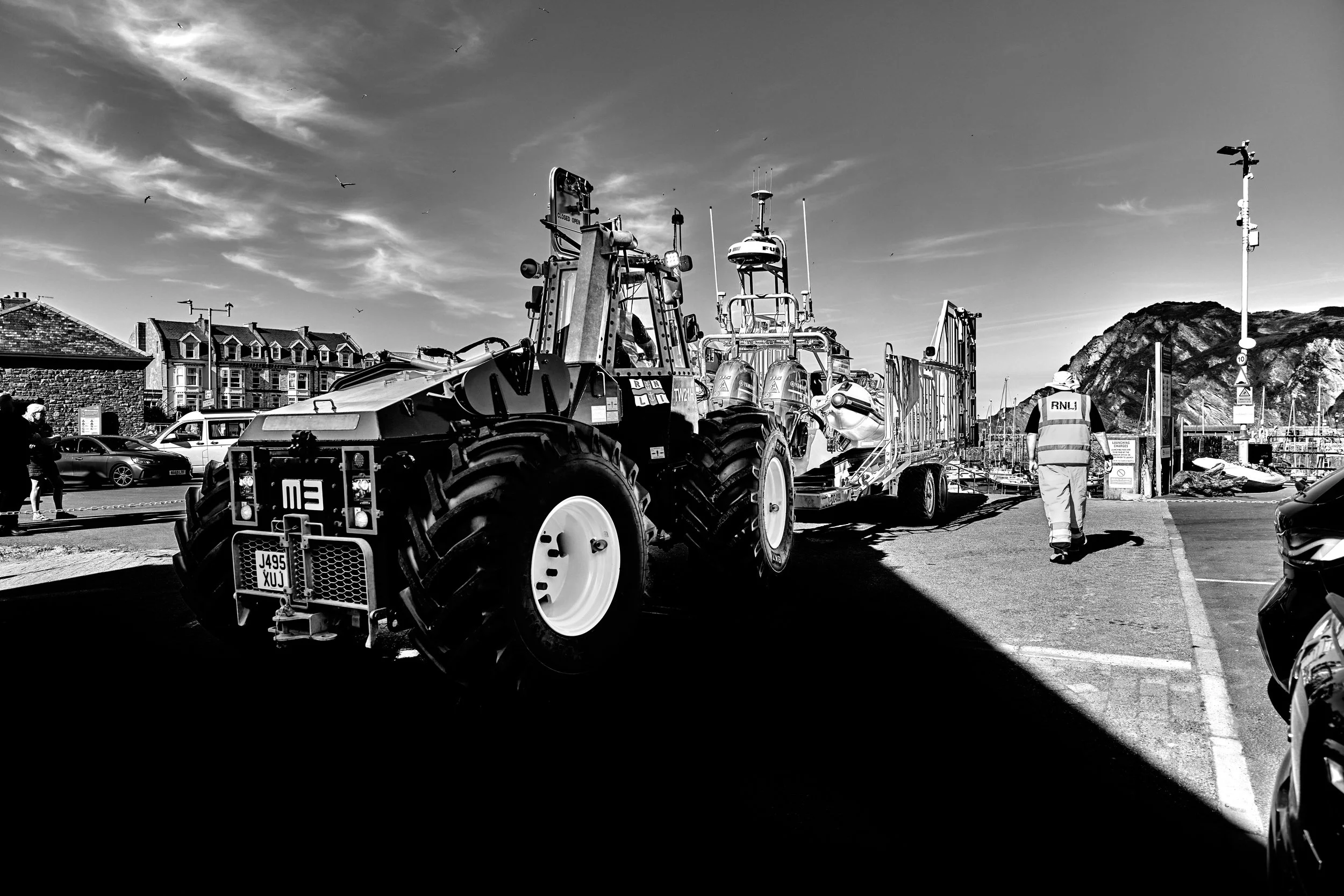 A black and white photo of a harbor with a large tractor and boat transportation trailer, a person in safety gear, boats docked in the background, and a cliff with buildings on top under a partly cloudy sky.