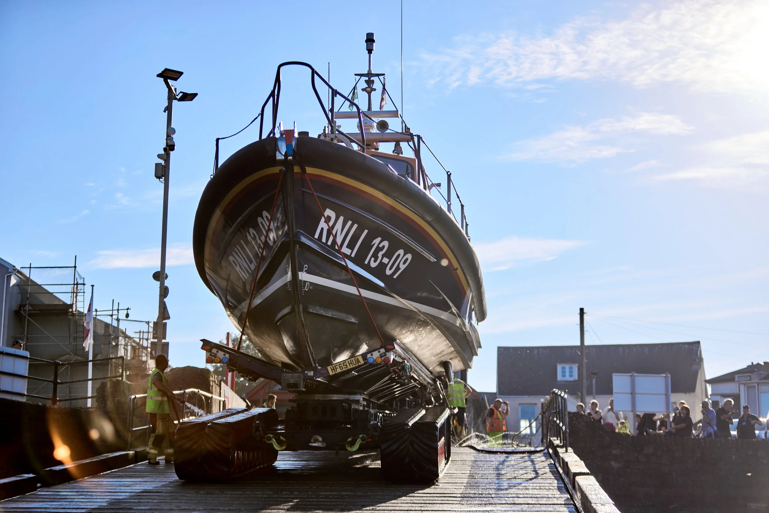 A large boat is being transported on a trailer, surrounded by construction workers and spectators under a bright blue sky.