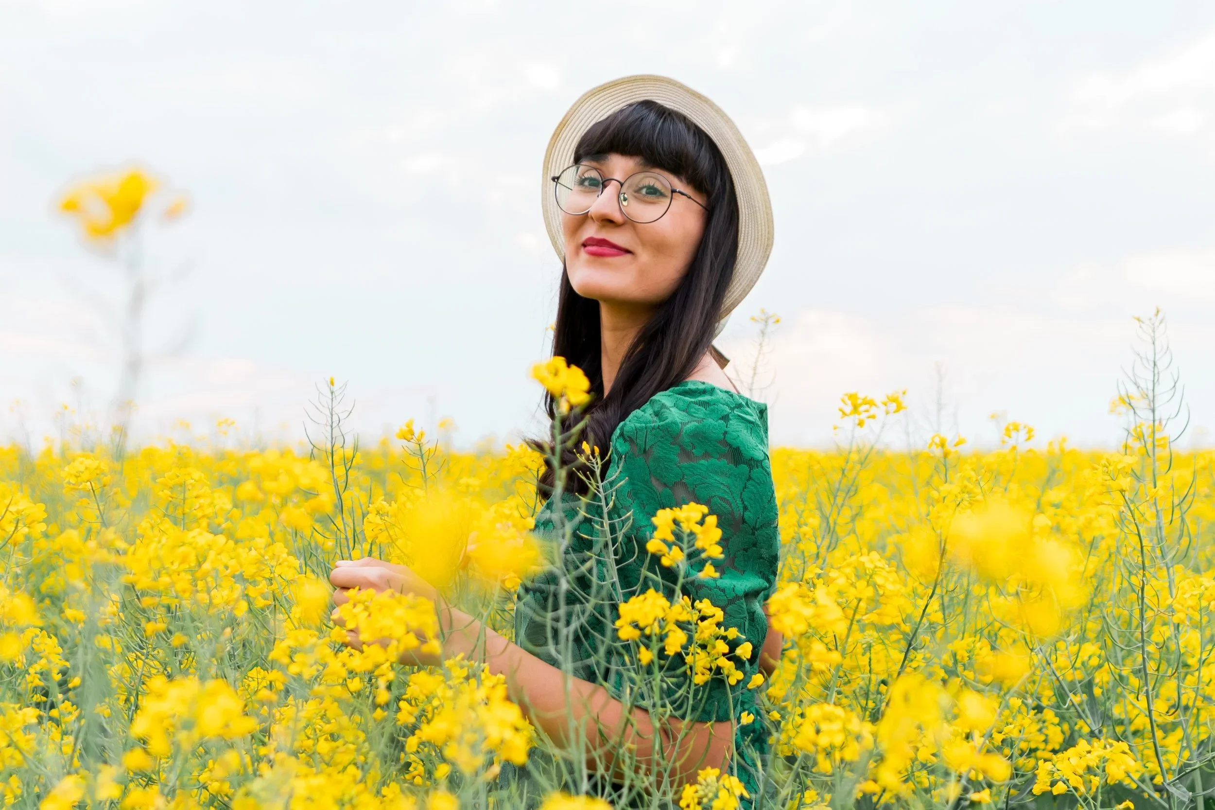 A woman with black hair, glasses, and a straw hat standing in a field of yellow flowers, wearing a green lace dress.