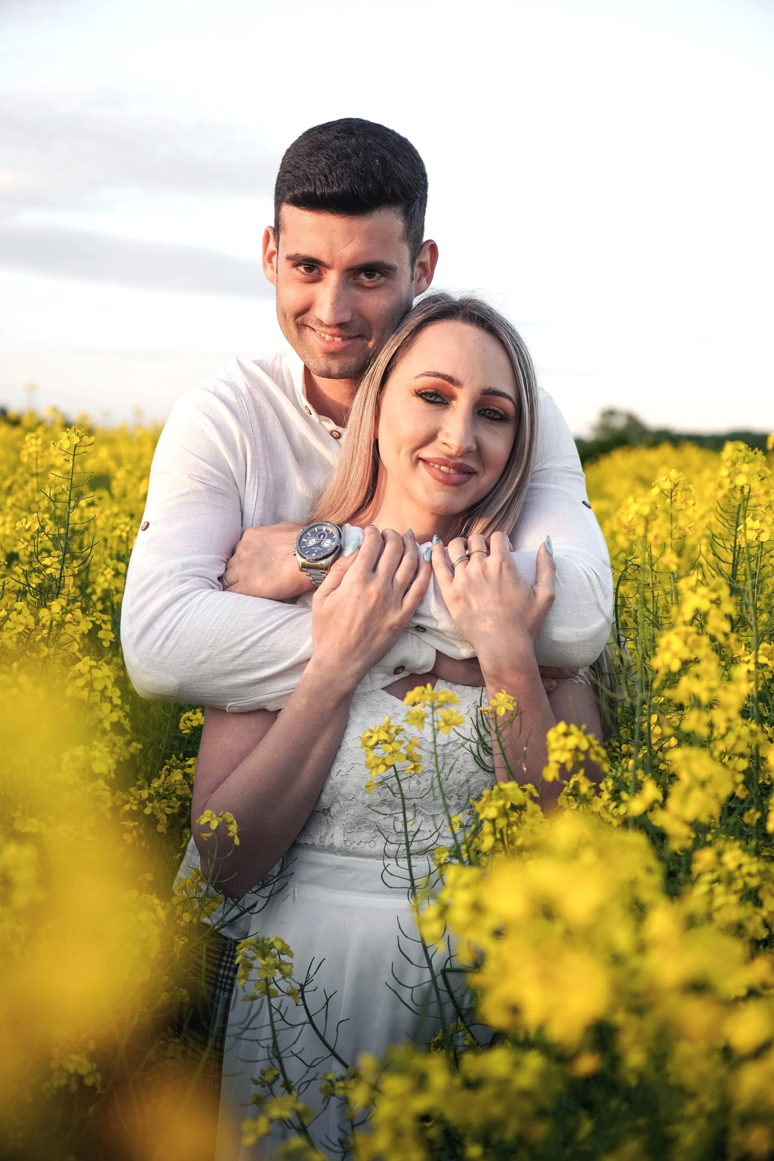 A smiling couple embracing in a yellow flower field during sunset, the man standing behind the woman with his arms around her shoulders. She wears a white dress, and he wears a white shirt. The woman has blonde hair, and the man has dark hair and a watch on his left wrist.