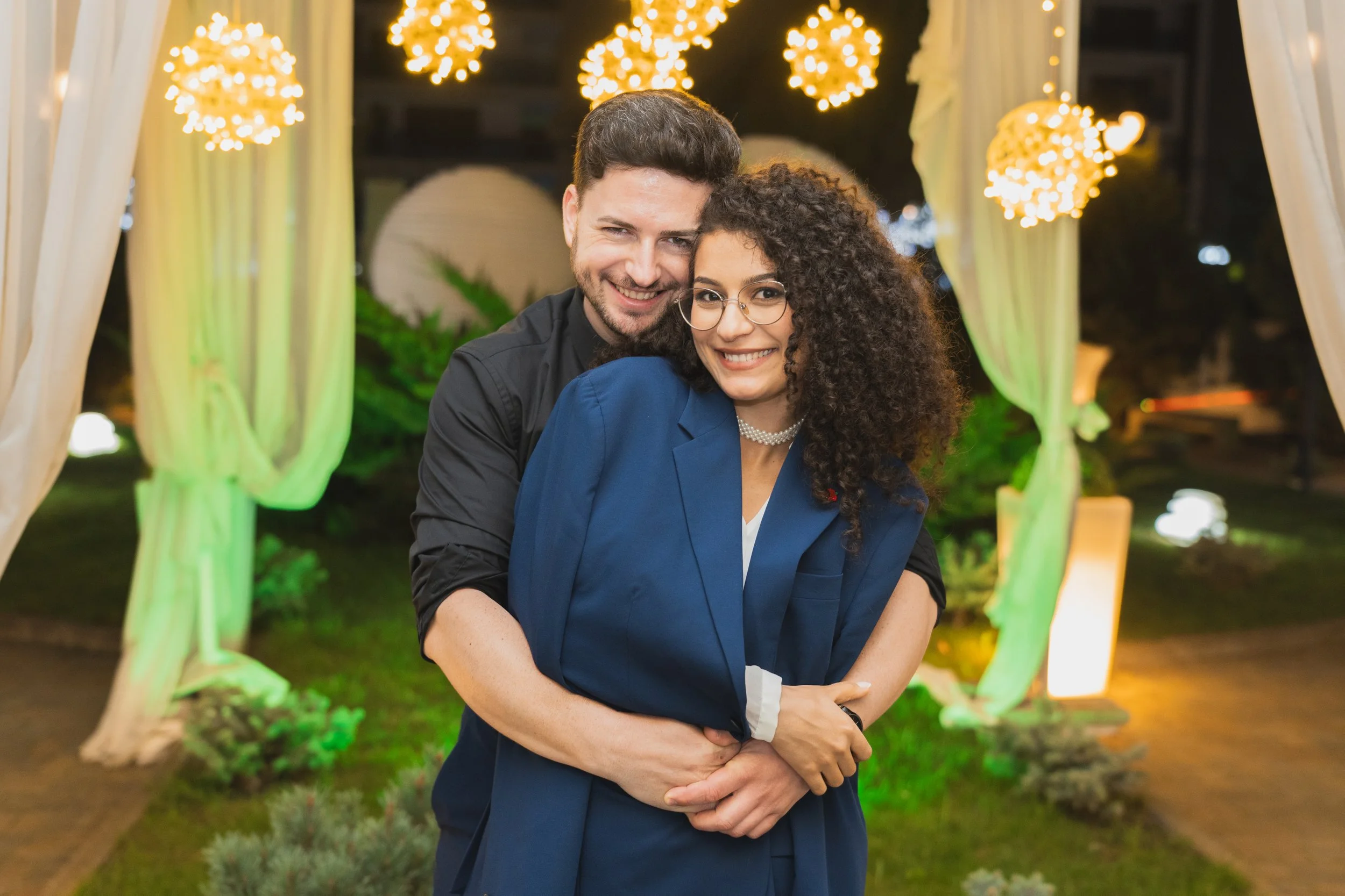 A happy couple embraces at night under decorated outdoor lights in a garden setting.