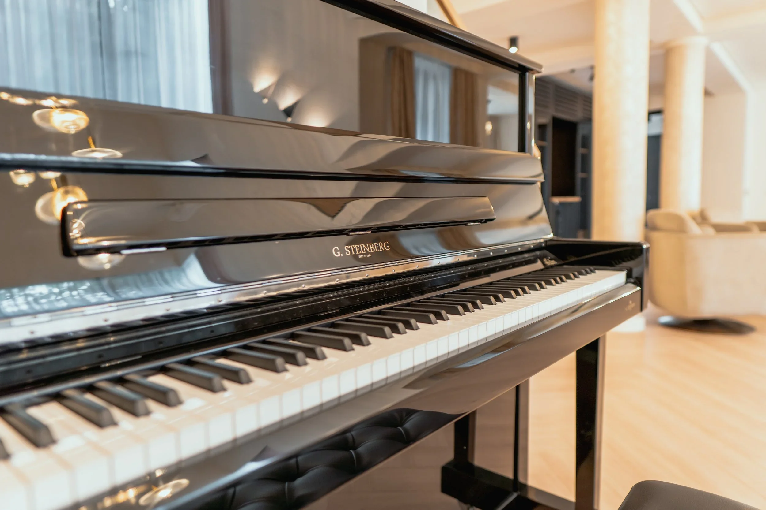 Close-up of a black grand piano in a well-lit lounge area with beige walls, columns, and a beige couch in the background.