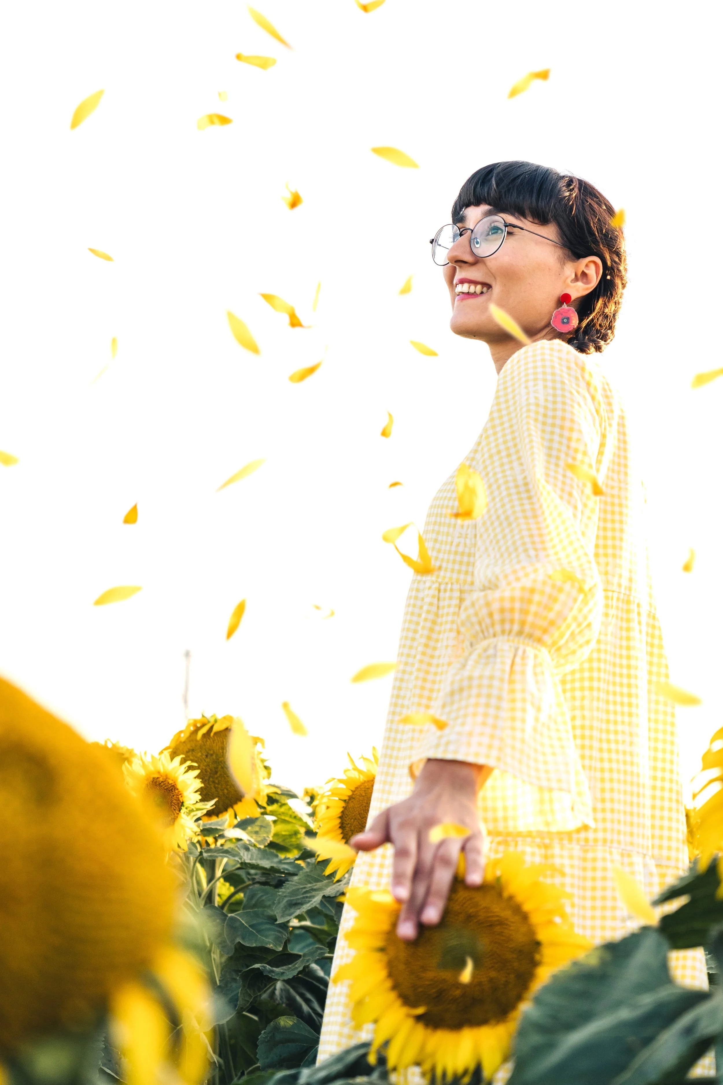 A woman with short dark hair, glasses, and red earrings smiling in a sunflower field, holding a sunflower and surrounded by falling yellow petals.