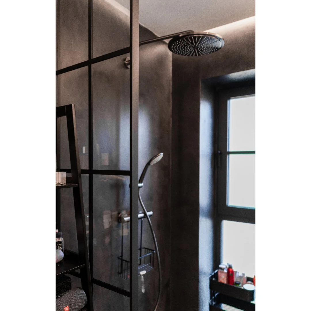 A modern shower area with a large round rain shower head attached to the ceiling, a handheld shower wand, a glass partition, a black shelf with toiletries, and a window providing natural light.