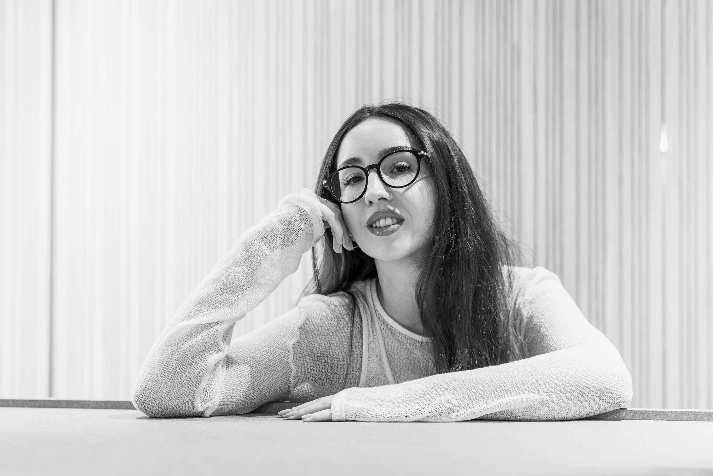 Black and white photo of a young woman with long hair, wearing glasses and a long-sleeve shirt, resting her head on her hand at a table in front of a vertically striped wall.