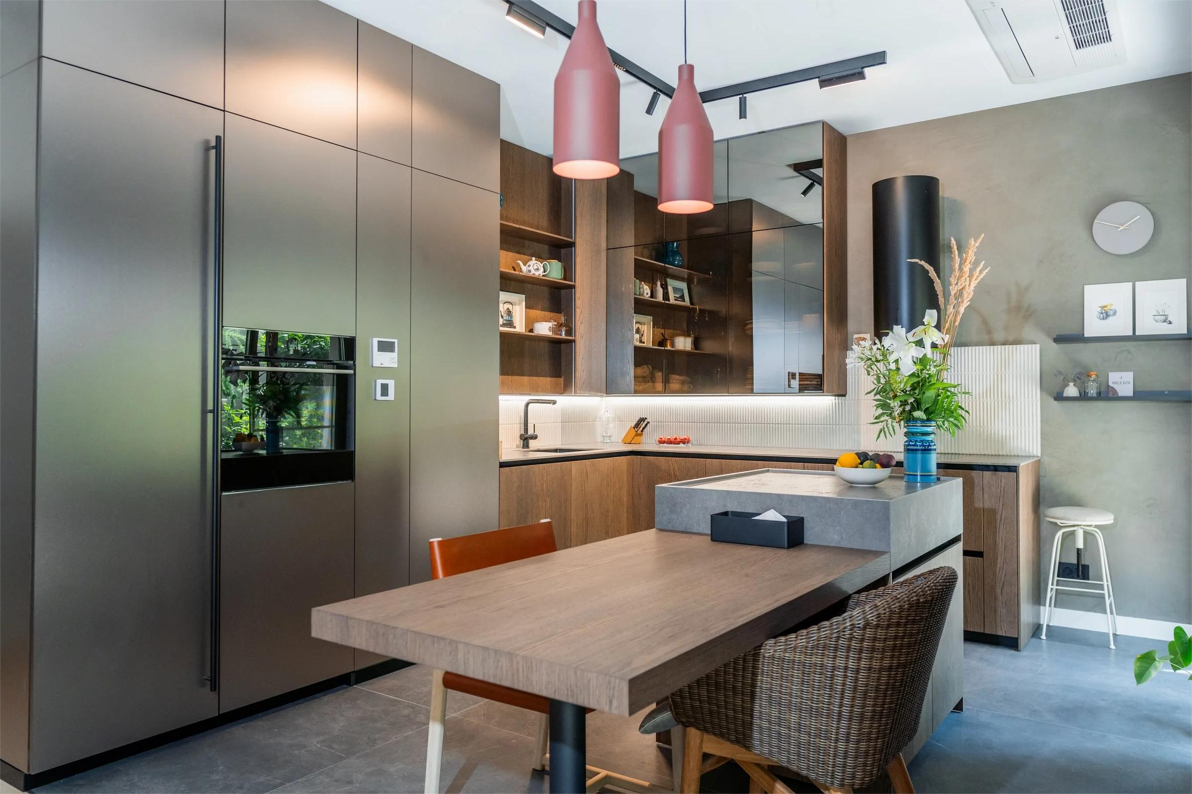Modern kitchen with stainless steel refrigerator, wooden cabinets, a gray island with a flower arrangement, pink pendant lights, and a green wall with floating shelves and artwork.