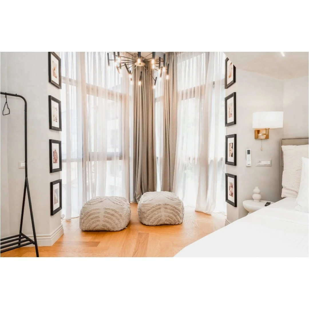 Bright hotel room with large window, sheer curtains, two pouf chairs, framed artwork, a bed, nightstand, and modern lighting fixtures.